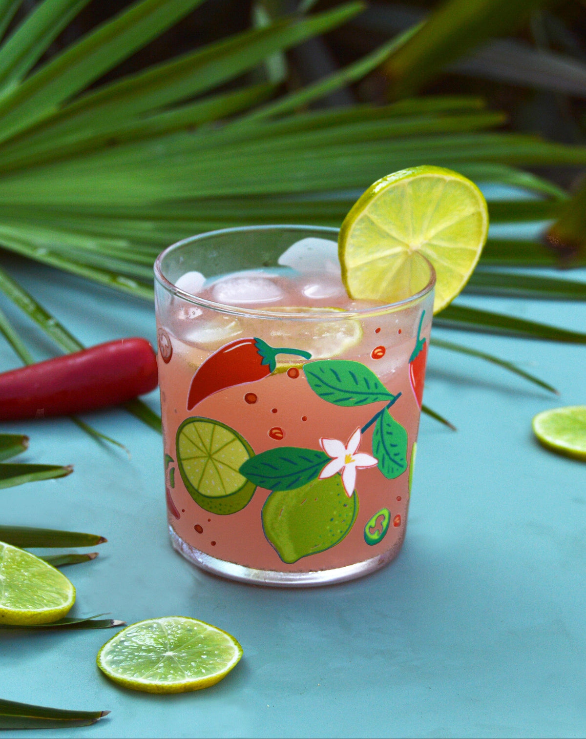 A clear glass tumbler with a colourful tropical fruit and flower print, filled with a pink drink and ice, garnished with a lime wheel, set on a blue surface with limes, palm leaves, and a red chili pepper in the background.